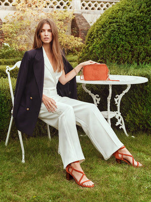 woman sitting on a garden chair wearing the heeled brancasters in sunset orange suede while holding the finsbury crossbody bag in sunset orange suede.