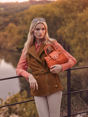 women wearing the primrose gilet in tan suede and holding the mini windsor in sunset orange suede proudly.