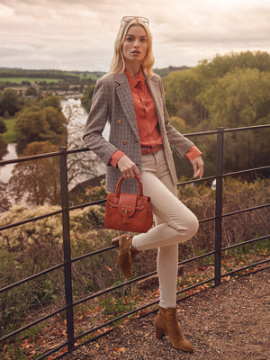 woman leaning against balcony overlooking a river and forest, wearing fairfax and favor sennowe belt and mini windsor crossbody bag in vibrant sunset orange. 